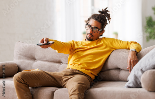 Bored tired african american man changing tv channel with remote control while relaxing at home