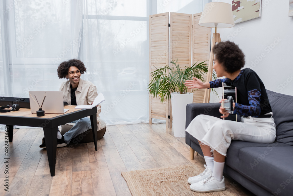 Smiling african american man looking at girlfriend with acoustic guitar near laptop and synthesizer at home. Smiling african american man looking at girlfriend with acoustic guitar near laptop and synthesizer at home.