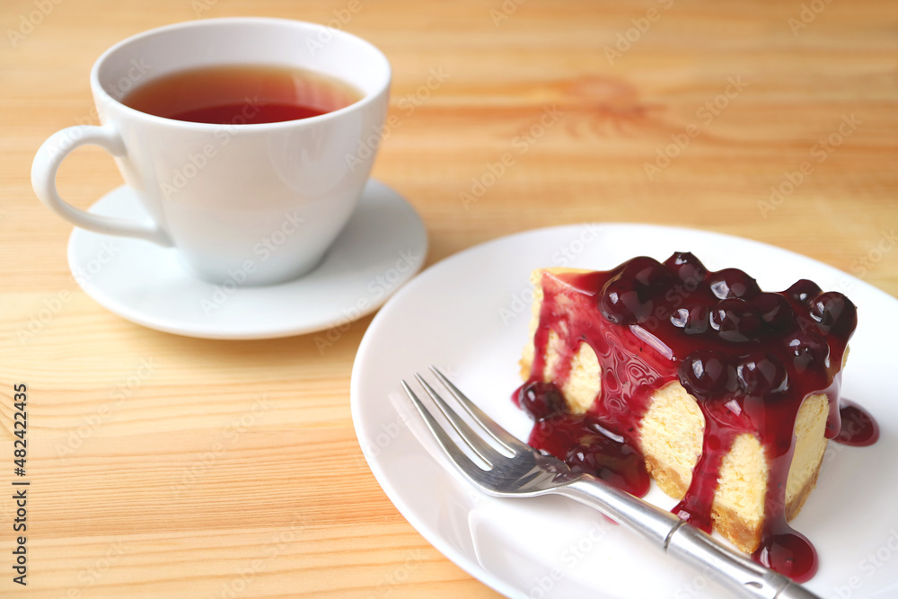 Plate of a slice of cheesecake with blueberry sauce and a cup of hot tea served on wooden table Plate of a slice of cheesecake with blueberry sauce and a cup of hot tea served on wooden table