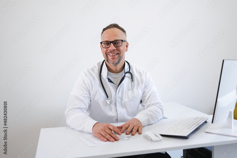 Portrait of smiling doctor in eyeglasses and in white coat sitting at his workplace at office Portrait of smiling doctor in eyeglasses and in white coat sitting at his workplace at office