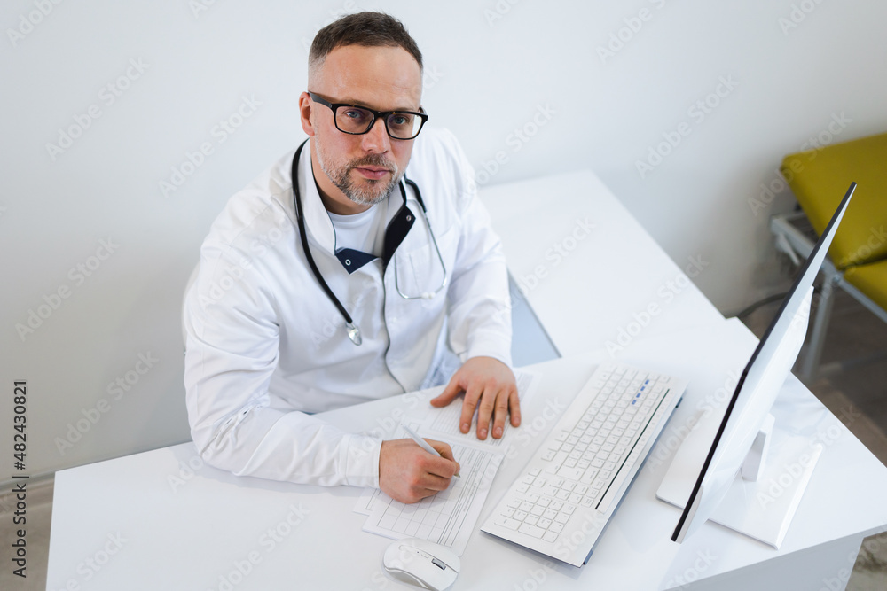 Male doctor filling medical documentation while sitting at his desk in the office Male doctor filling medical documentation while sitting at his desk in the office