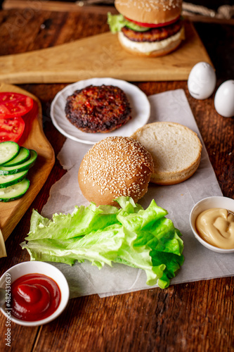 Cooking hamburger or cheeseburger. Different ingredients for a classic hamburger. Grilled meat, vegetables, greens, sauces near a sesame bun. Wooden background.