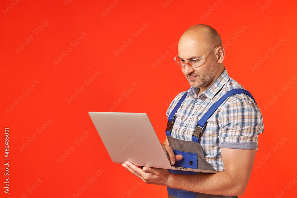Bald man in work clothes and protective glasses is working at a laptop holding it in his hands, a photo on a red background. The main worker looks at the project plan. Copy space Bald man in work clothes and protective glasses is working at a laptop holding it in his hands, a photo on a red background. The main worker looks at the project plan. Copy space