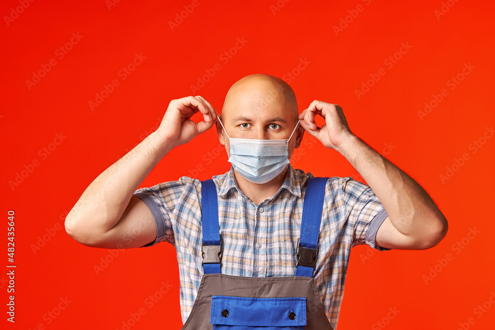 Bald man in work clothes puts on a mask and looks into the camera, a photo on a red background. The employee came to repair the equipment in a mask. The coronavirus epidemic. Bald man in work clothes puts on a mask and looks into the camera, a photo on a red background. The employee came to repair the equipment in a mask. The coronavirus epidemic.