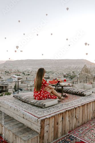 A young female traveller in a red dress is having early morning breakfast and watching hot air balloons in Cappadocia, Turkey on a hot summer day