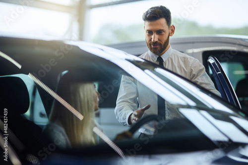 Caucasian salesman showing to female customer new luxury car at showroom. Happy young woman choosing new vehicle at auto salon. Purchase and people concept.