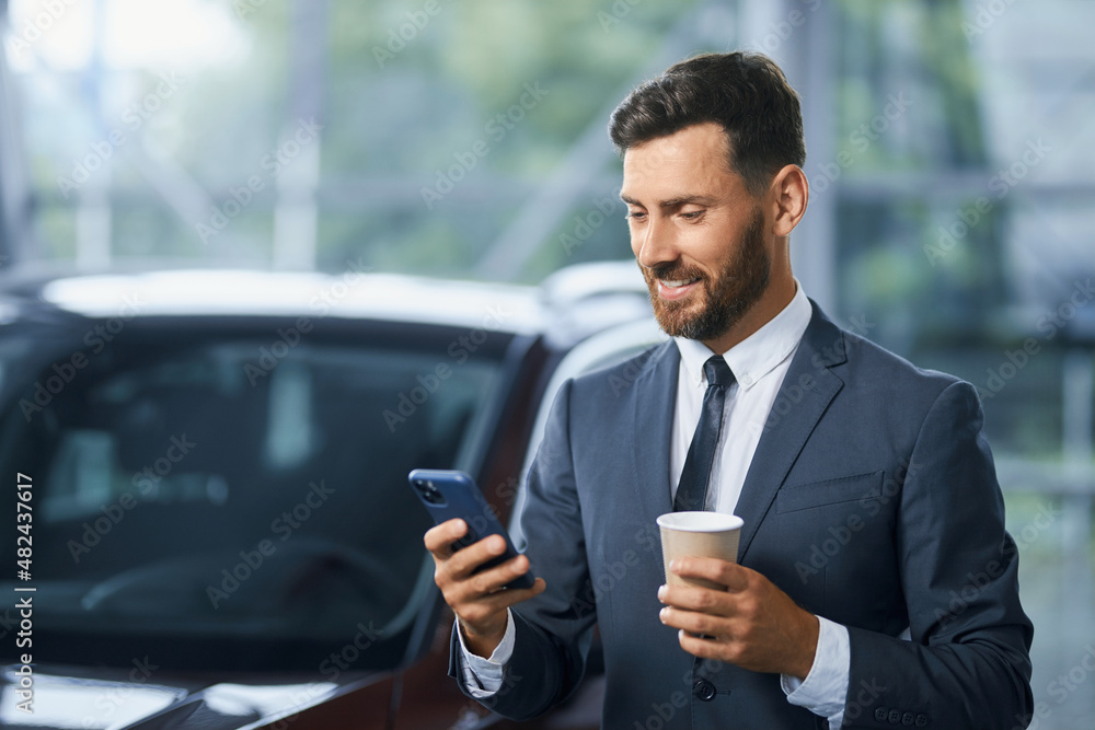 Handsome caucasian businessman drinking coffee and using modern smartphone while buying new car at salon. Concept of people, technology and transport. Handsome caucasian businessman drinking coffee and using modern smartphone while buying new car at salon. Concept of people, technology and transport.