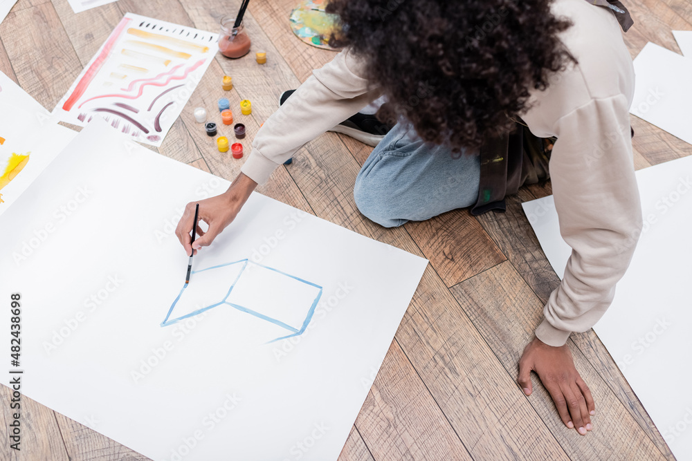 Overhead view of african american man painting on paper near paints on floor at home. Overhead view of african american man painting on paper near paints on floor at home.