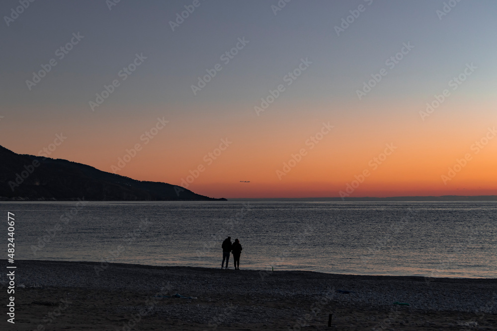 Relationship, Valentine's day, romance concept photo. Silhouettes of a couple by the sea. Beautiful red, orange sunset. Sand, sea and mountain view. Horizontal web banner. 