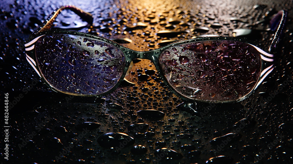 Women's sunglasses in water drops on a wet surface in the dark colorful neon lights, abstraction, selective focus
