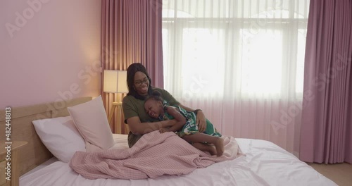 Loving African American mom hugging soothing an adorable, sweet baby girl lying in bed, cheerful family small kid girl and mom embrace cuddle laugh relax in the bedroom.
