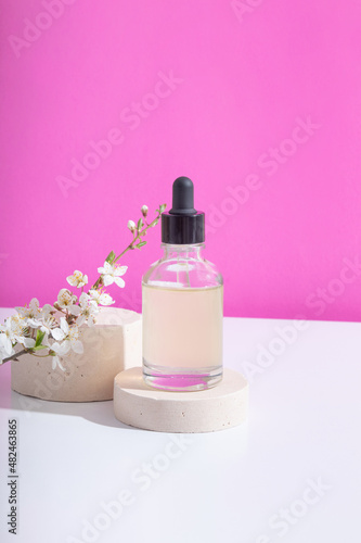 Glass dropper bottle with serum on podium with blooming twigs on pink background. Vertical stock photo