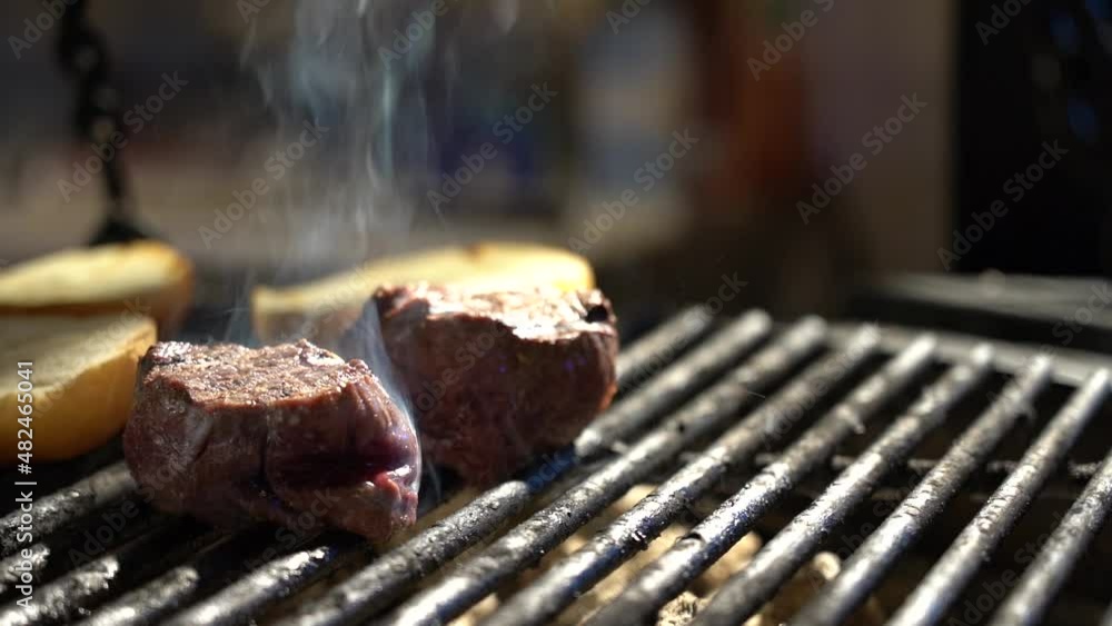 Filet mignon is fried on the grill close-up. In the background, cooking tongs are flipping a white bun. Filet mignon is fried on the grill close-up. In the background, cooking tongs are flipping a white bun.