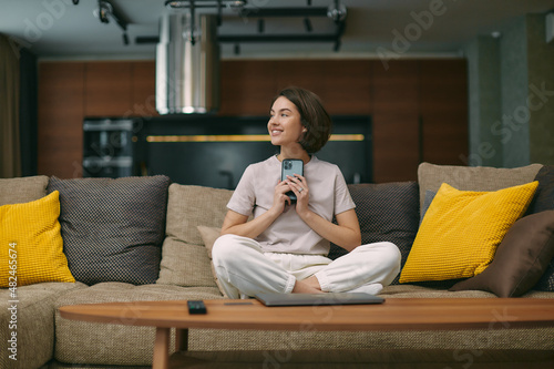 Dreaming inspired young girl sitting on couch, holding phone, receive pleasant message, looking to aside, smiling