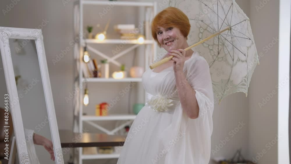Portrait of happy Caucasian senior redhead bride in elegant white dress posing with sun umbrella in slow motion indoors. Excited beautiful confident woman looking at camera smiling. Marriage concept Portrait of happy Caucasian senior redhead bride in elegant white dress posing with sun umbrella in slow motion indoors. Excited beautiful confident woman looking at camera smiling. Marriage concept