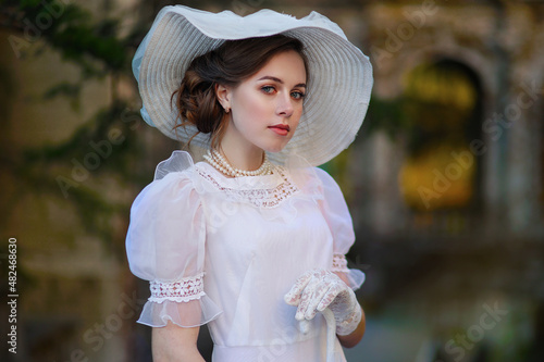 Fotografie Young woman in white edwardian style dress and white big hat standing in old abandoned castle