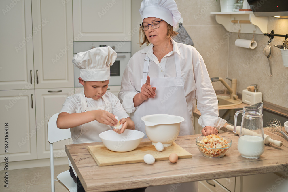 Mother and son cooking apple pie in the home kitchen. A woman and a boy in chef hats and aprons cook with pastries Mother and son cooking apple pie in the home kitchen. A woman and a boy in chef hats and aprons cook with pastries