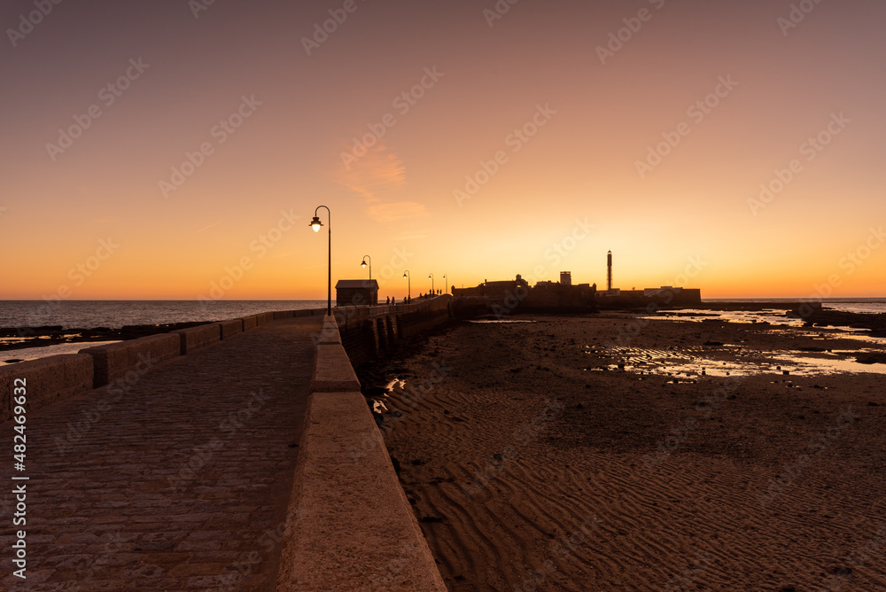 Pedestrian promenade to an islet with the lighthouse and San Sebastian Castle in the old town near La Caleta beach at sunset, Cadiz, Andalusia, Spain