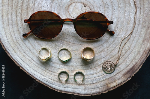 Wooden tray with round tortoiseshell sunglasses, gold necklace with pendant, hoop earrings and various rings. Flat lay.