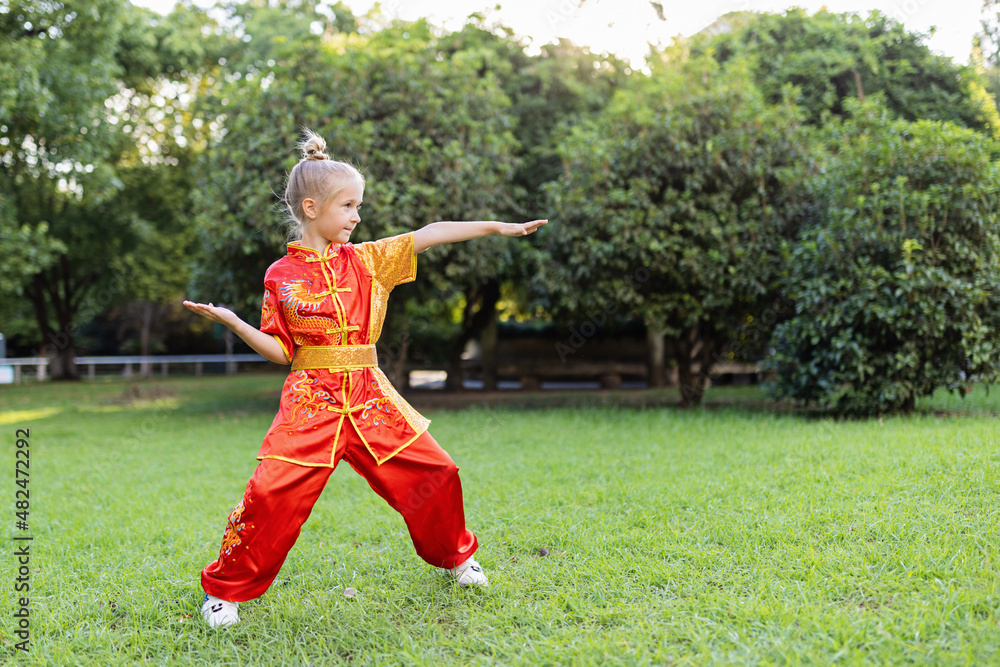 Fototapeta premium Cute little caucasian girl seven years old in red sport wushu uniform exercising in park at summer day. Lifestyle portrait of kung fu fighter child athlete