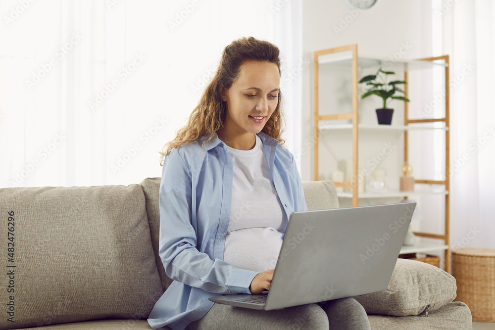 Pregnant woman sitting on comfortable sofa in living room, working on laptop computer, reading parenthood information, ordering newborn baby clothes, having virtual telemedicine pregnancy consultation Pregnant woman sitting on comfortable sofa in living room, working on laptop computer, reading parenthood information, ordering newborn baby clothes, having virtual telemedicine pregnancy consultation