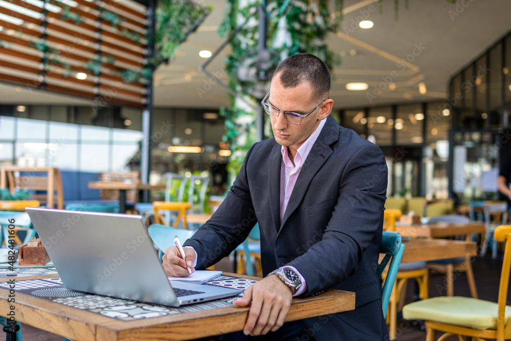 Businessman working on his laptop and writing in a notebook while sitting in a cafe Businessman working on his laptop and writing in a notebook while sitting in a cafe
