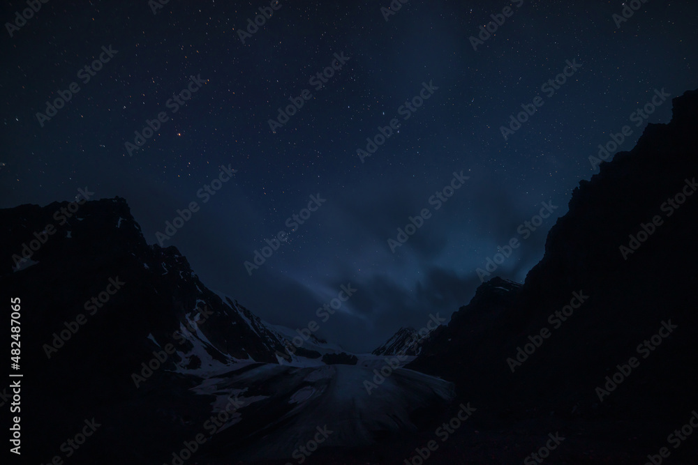 Awesome mountain landscape with large glacier tongue and black silhouettes of rocks under clouds in night starry sky. Atmospheric alpine scenery with glacier and mountains silhouettes in starry night.