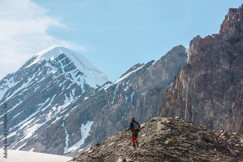 Scenic alpine landscape with hiker with trekking poles against snow mountain peak and sharp rocks in sunlight. Man with backpack walks in high mountains under cirrus clouds in blue sky in sunny day.