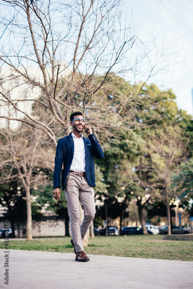 Wide shot smiling black man entrepreneur walking through a park while using his phone doing business