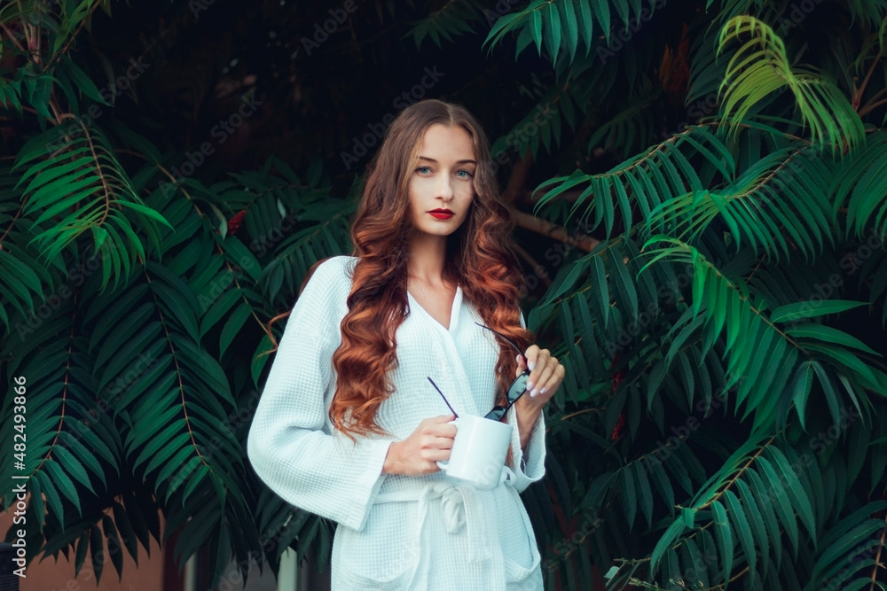 Gorgeous girl in a white coat against the background of large green leaves. Cup and black glasses in hands. Red lipstick and arrows. Morning portrait Gorgeous girl in a white coat against the background of large green leaves. Cup and black glasses in hands. Red lipstick and arrows. Morning portrait