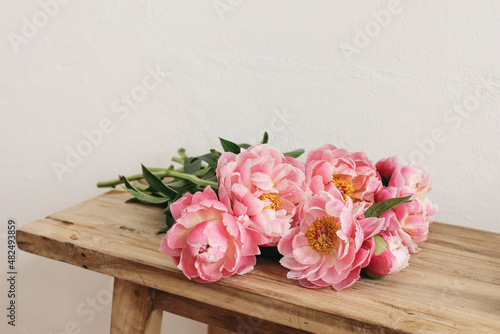 Fototapeta Naklejka Na Ścianę i Meble -  Blooming pink peonies flowers, bouquet lying on old wooden table, bench. White wall. Selective focus, blurred background. Wedding or birthday celebration, gift concept. Floral still life.