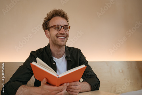 Portrait of smiling university student studying, learning language, exam preparation at library, education concept. Emotional handsome man wearing stylish eyeglasses reading book looking away 