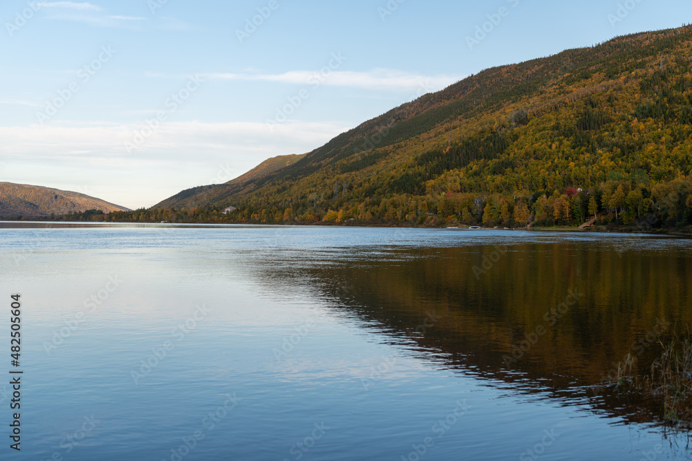 A wide river with smooth blue water. There is a luscious green, red and golden forest of trees along the riverbank.  The calm and peaceful pond water is reflecting the sky and autumn landscape.