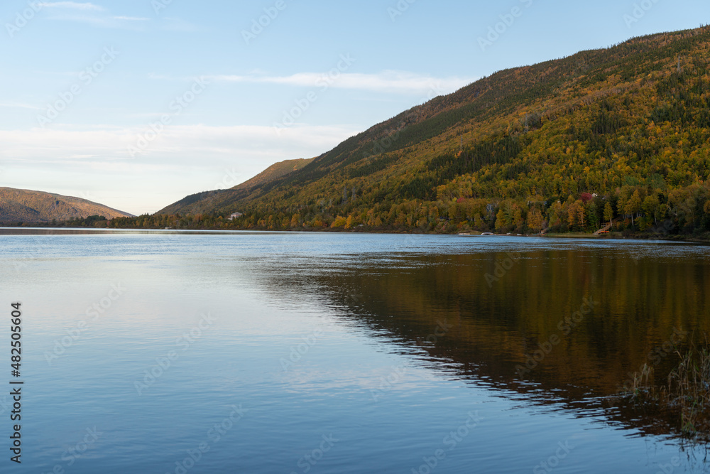 Fototapeta premium A wide river with smooth blue water. There is a luscious green, red and golden forest of trees along the riverbank. The calm and peaceful pond water is reflecting the sky and autumn landscape.