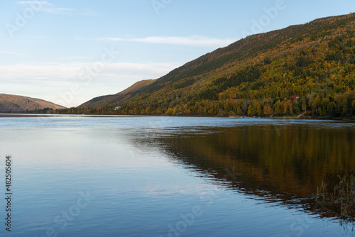 A wide river with smooth blue water. There is a luscious green, red and golden forest of trees along the riverbank. The calm and peaceful pond water is reflecting the sky and autumn landscape.