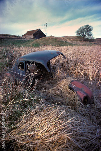 Derilect car body in field of weeds with barn and windmill in background. photo
