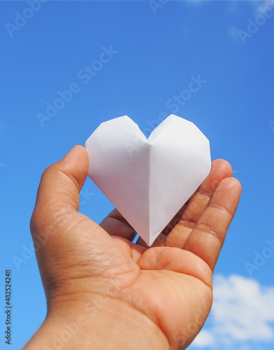 Hand holding a white paper heart on the sky background.The heart is a symbol of love. and white represents purity.