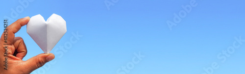 Hand holding a white paper heart on the sky background.The heart is a symbol of love. and white represents purity.