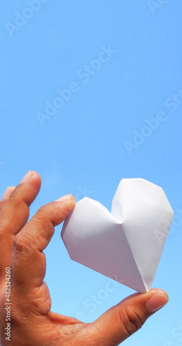 Hand holding a white paper heart on the sky background.The heart is a symbol of love. and white represents purity.