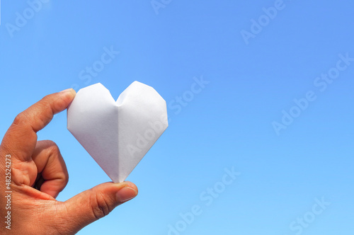 Hand holding a white paper heart on the sky background.The heart is a symbol of love. and white represents purity.