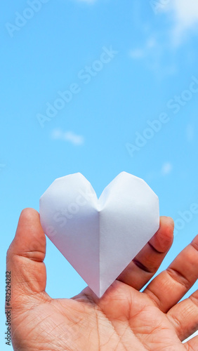 Hand holding a white paper heart on the sky background.The heart is a symbol of love. and white represents purity.