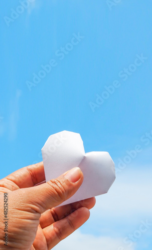Hand holding a white paper heart on the sky background.The heart is a symbol of love. and white represents purity.