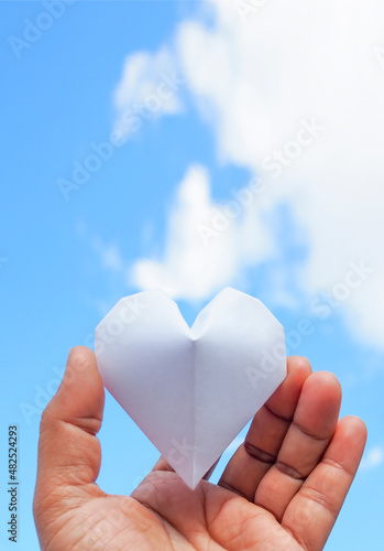 Hand holding a white paper heart on the sky background.The heart is a symbol of love. and white represents purity.