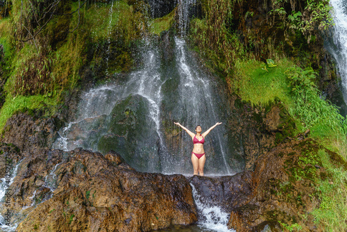 Full view of caucasian woman in swimsuit at thermal springs in Colombia. Horizontal panoramic view of woman raising up arms underneath a big waterfall in the jungle. Colombia travel destination photo