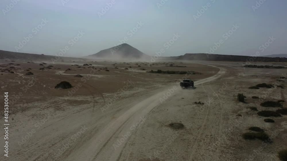 Aerial View Of Dust Trail From 4x4 Driving Across Arid Desert Landscape With Mud Volcano In Background. Tracking Aerial View Of Dust Trail From 4x4 Driving Across Arid Desert Landscape With Mud Volcano In Background. Tracking