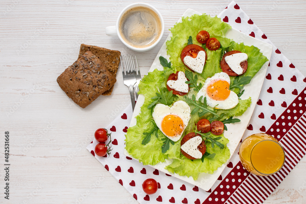 Gala breakfast for two on Valentine's Day. A plate of heart-shaped fried eggs, tomatoes, cheese, coffee and juice on a white wooden table covered with a napkin. View from above. Space for text. Gala breakfast for two on Valentine's Day. A plate of heart-shaped fried eggs, tomatoes, cheese, coffee and juice on a white wooden table covered with a napkin. View from above. Space for text.
