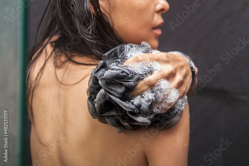 Obraz na plátně A mixed race female is washing her back with a charcoal soapy loofah in a black