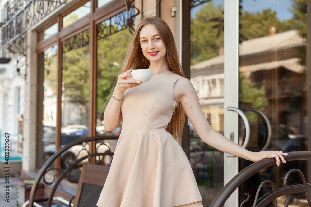 Beautiful woman in a dress stands on the porch of cafe with coffee cup