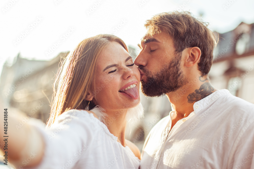 Selfie portrait of a young couple in love. Man kisses his girlfriend on cheek in city. Having fun Selfie portrait of a young couple in love. Man kisses his girlfriend on cheek in city. Having fun