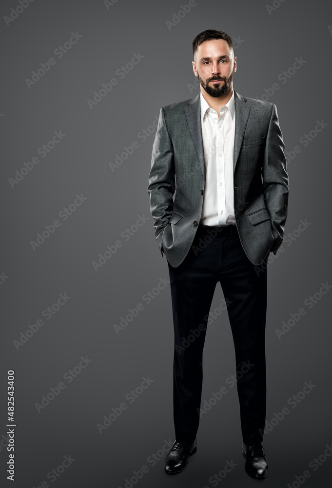 Fashion Handsome Bearded Man in Gray Suit and White Shirt without Tie. Elegant Businessman in Open Collar Shirt, Hands in Pocket Full Length over Gray Background Fashion Handsome Bearded Man in Gray Suit and White Shirt without Tie. Elegant Businessman in Open Collar Shirt, Hands in Pocket Full Length over Gray Background